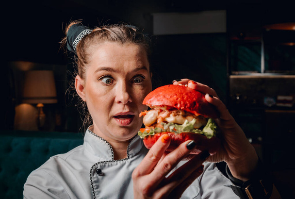 young woman chef with burger in hands on dark background in cafe - Фото, зображення