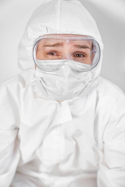 portrait of a tired doctor in protective suit, glasses, mask against white wall - Фото, зображення