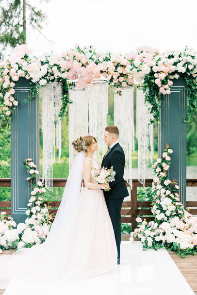 Beautiful caucasian Wedding couple is standing in front of decorated with pink roses archway and holding hands, wedding ceremony, wedding vows - Foto, Imagem
