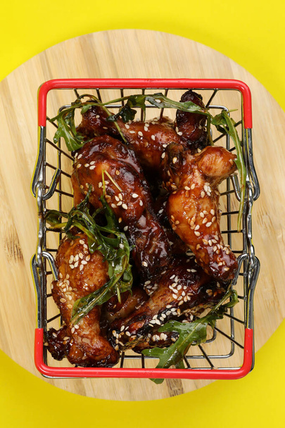 Top view of a fresh cooked delicious chicken wings with green arugula in a metal shopping basket isolated on a yellow background. Food delivery. - Photo, Image