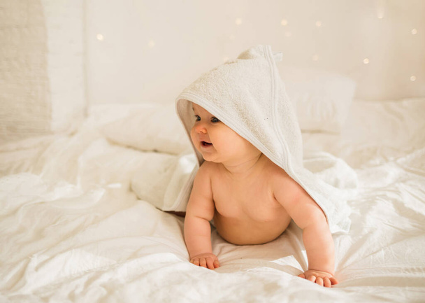little baby girl in a white towel with a hood is on all fours on a white blanket on the bed - Photo, Image