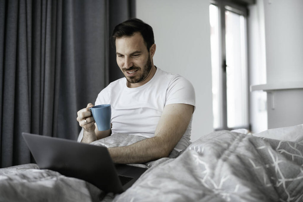 Mladý muž sleduje film v posteli a pije kávu. Hezký muž si užívá ráno. - Fotografie, Obrázek