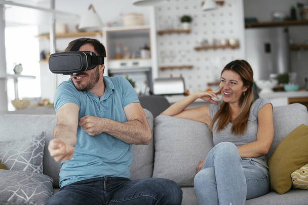 Happy couple having fun with virtual reality glasses in living room. Young couple playing and enjoying video games with VR headset at home. - 写真・画像