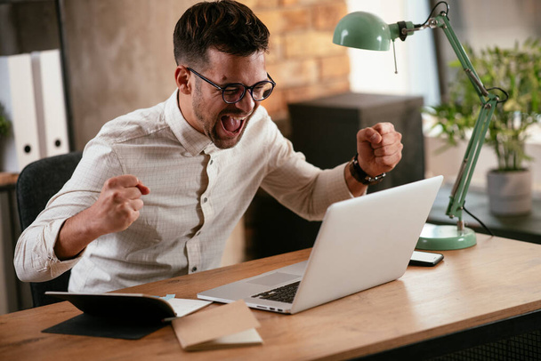 Szczęśliwy młody biznesmen krzyczy podczas pracy w biurze na swoim laptopie. - Zdjęcie, obraz