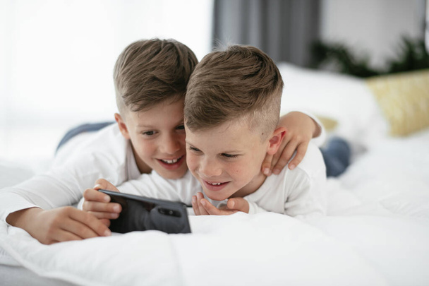 Two little brothers are playing games on the phone. Beautiful boys enjoying at home. - Photo, Image