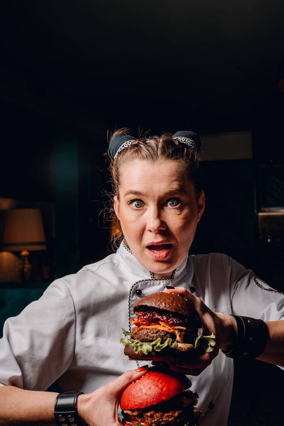 mujer joven chef con dos hamburguesas en las manos sobre fondo oscuro en la cafetería - Foto, imagen