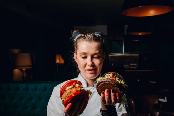молодая женщина шеф-повар с двумя бургерами в руках на темном фоне в кафе - Фото, изображение