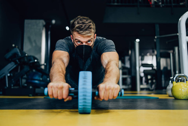 Good looking handsome male athlete with protective face mask exercising in modern fitness gym. Dark muddy light with strong shadows. Pandemic sports indoors concept. - Photo, image