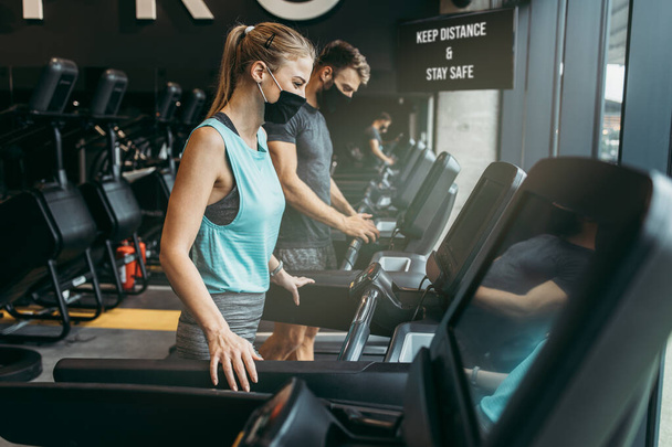 Young fit woman and man running on treadmill in modern fitness gym. They keeping distance and wearing protective face masks. Coronavirus world pandemic and sport theme. - Фото, изображение