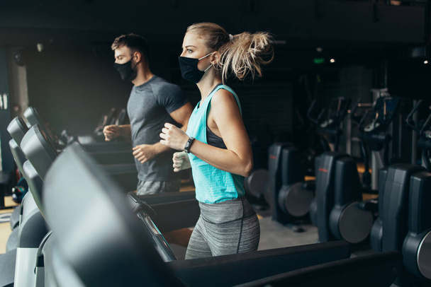 Young fit woman and man running on treadmill in modern fitness gym. They keeping distance and wearing protective face masks. Coronavirus world pandemic and sport theme. - Φωτογραφία, εικόνα