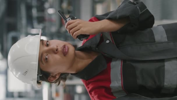 Verticale slow mo portret van geschoolde gemengde ras vrouwelijke fabrieksarbeider met walkie-talkie kijken naar camera op de werkplek - Video