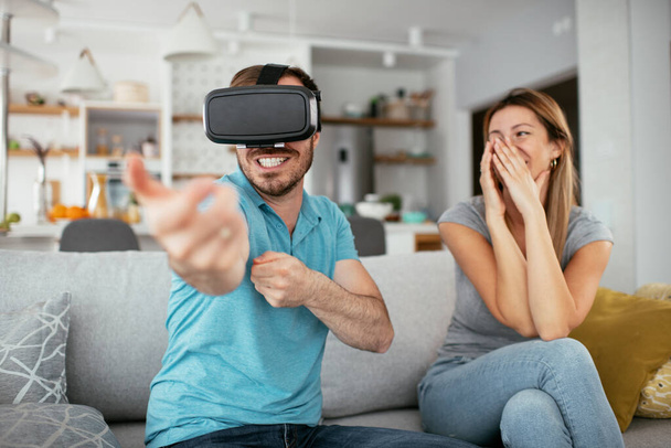 Happy couple having fun with virtual reality glasses in living room. Young couple playing and enjoying video games with VR head sets at home. - Fotó, kép