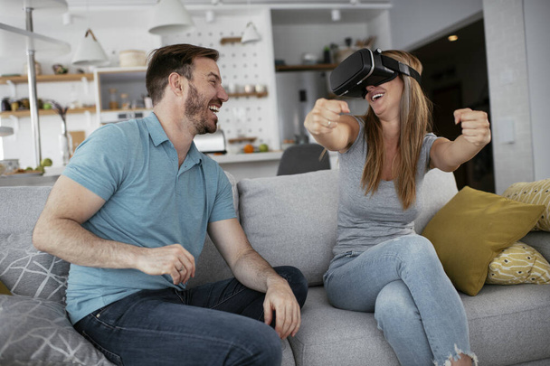 Счастливая пара веселится с очками виртуальной реальности в гостиной. Молодая пара, играющая и наслаждающаяся видеоиграми с VR гарнитурой дома. - Фото, изображение