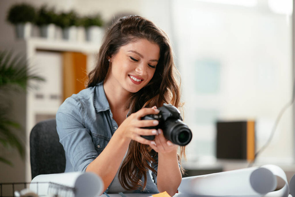Happy young female executive looking at camera in creative office. Freelance photographer woman with camera at home office - Foto, afbeelding