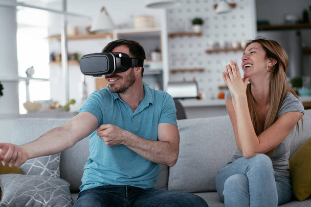 Счастливая пара веселится с очками виртуальной реальности в гостиной. Молодая пара, играющая и наслаждающаяся видеоиграми с VR гарнитурой дома. - Фото, изображение