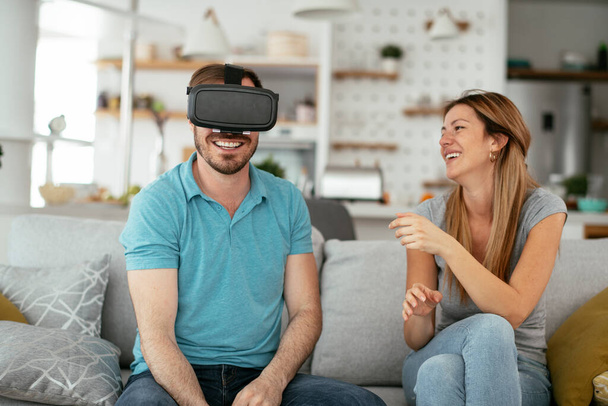 Casal feliz se divertindo com óculos de realidade virtual na sala de estar. Casal jovem jogando e desfrutando de jogos de vídeo com fone de ouvido VR em casa. - Foto, Imagem