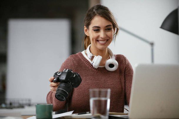 Счастливая молодая исполнительница смотрит в камеру в креативном кабинете. внештатный фотограф женщина с камерой на домашнем офисе редактирования фотографий на компьютере - Фото, изображение