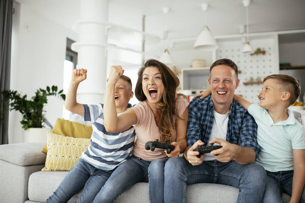 Πατέρας και μητέρα διασκεδάζουν παίζοντας βιντεοπαιχνίδια με παιδιά στο σπίτι. - Φωτογραφία, εικόνα