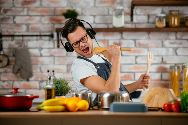 Mutfakta yemek pişirirken müzik dinleyip dans eden kulaklı mutlu bir adam.. - Fotoğraf, Görsel