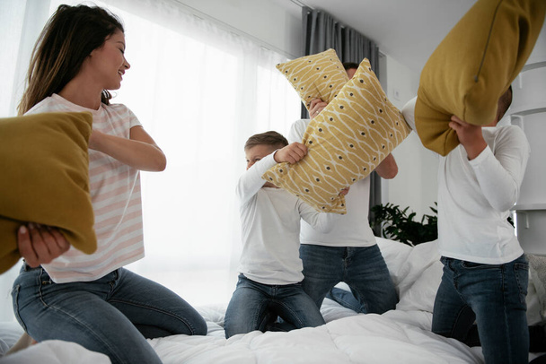 Jovens pais com filhos lutando almofadas na cama. Mãe feliz e pai com meninos se divertindo em casa. - Foto, Imagem
