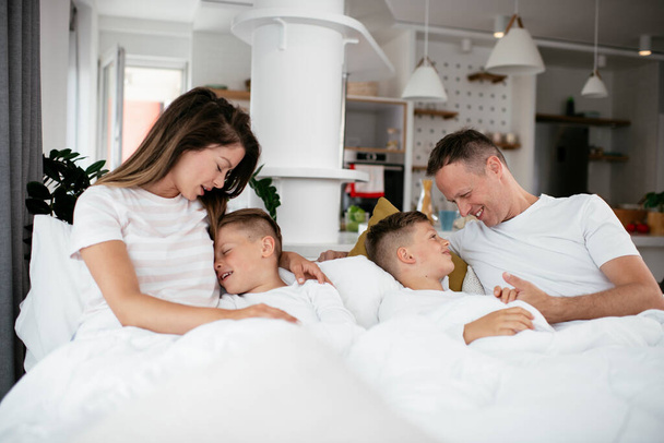 Nuori perhe nauttii sängyssä. Onnelliset vanhemmat poikien kanssa rentouttava sängyssä. - Valokuva, kuva