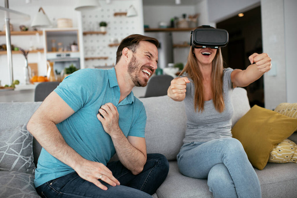 Szczęśliwa para bawiąca się okularami wirtualnej rzeczywistości w salonie. Młoda para grająca i ciesząca się grami wideo ze słuchawkami VR w domu. - Zdjęcie, obraz