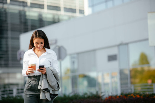 Schöne Frau geht mit Handy in der Hand zur Arbeit, wenn sie in der Nähe von Bürogebäuden spaziert. - Foto, Bild