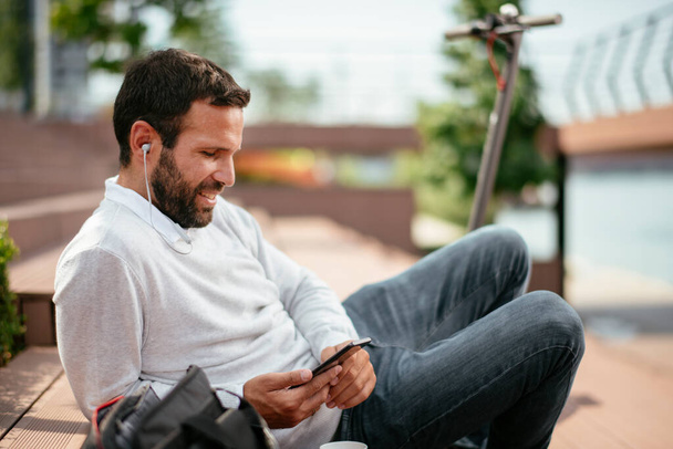 Portret przystojnego biznesmena na świeżym powietrzu. Młody człowiek ze słuchawkami siedzi na zewnątrz i używa telefonu. - Zdjęcie, obraz