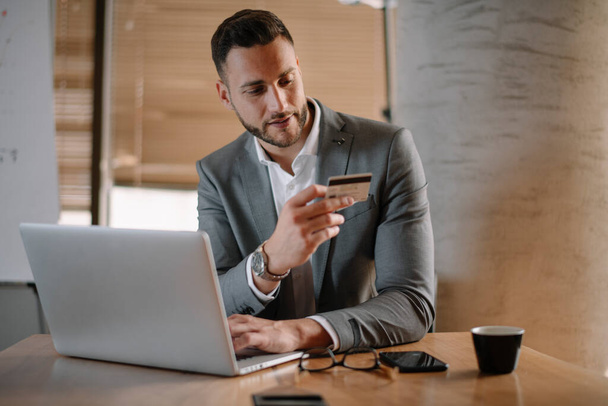 Przystojny biznesmen w klasycznym garniturze z laptopem, trzymający kartę kredytową na zakupy online. - Zdjęcie, obraz