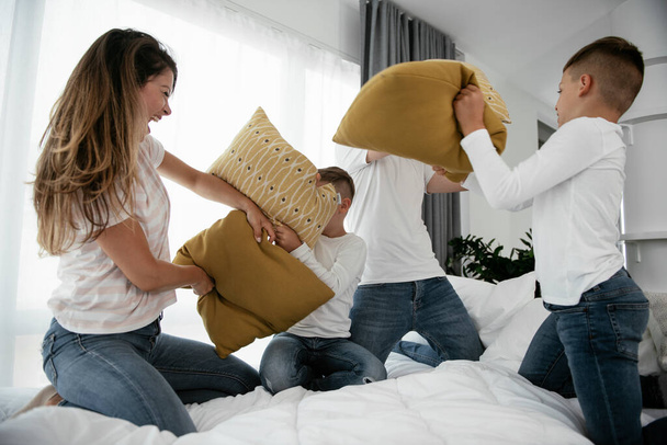 Νέοι γονείς με γιους που παλεύουν με μαξιλάρια στο κρεβάτι. Ευτυχισμένοι γονείς με αγόρια που διασκεδάζουν στο σπίτι. - Φωτογραφία, εικόνα