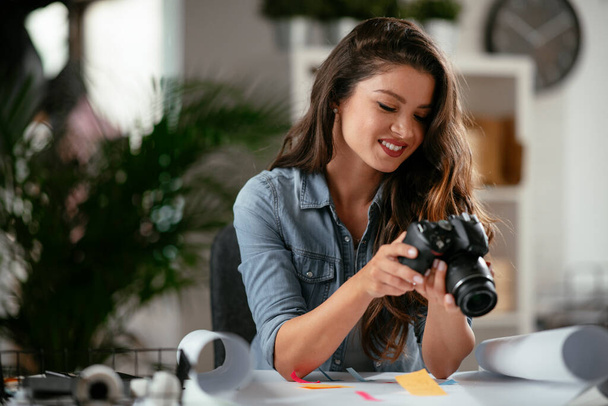 Χαρούμενη νεαρή γυναίκα στέλεχος που κοιτάζει την κάμερα στο δημιουργικό γραφείο. Ανεξάρτητη φωτογράφος γυναίκα με κάμερα στο γραφείο στο σπίτι - Φωτογραφία, εικόνα