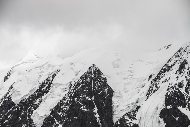 Атмосферный минималистский альпийский ландшафт с массивным висящим ледником на снежной вершине горы. Большой балкон на ледниковом краю. Низкие облака среди заснеженных гор. Величественные пейзажи на большой высоте. - Фото, изображение