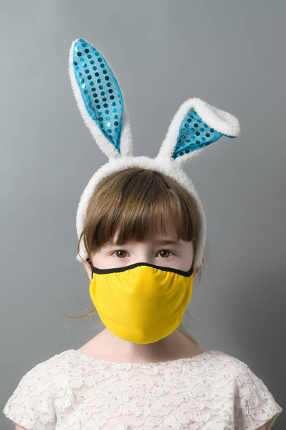 Aranyos kislány nyuszifül fejpánt és sárga védő maszk szürke stúdió háttér. Covid húsvét koncepció. Függőleges irány - Fotó, kép