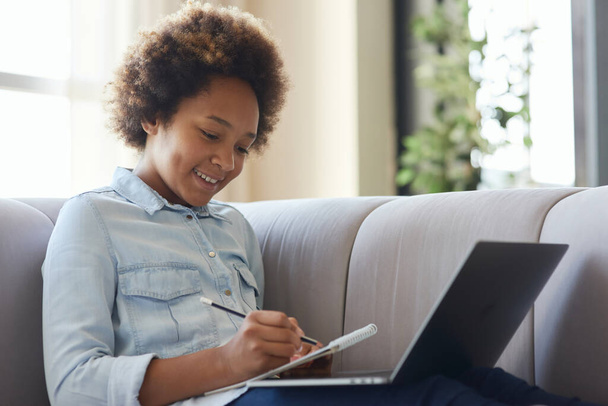 Explore and discover. Joyful mixed race teen schoolgirl using pencil for making notes while doing homework, sitting with laptop on the couch at home - Φωτογραφία, εικόνα