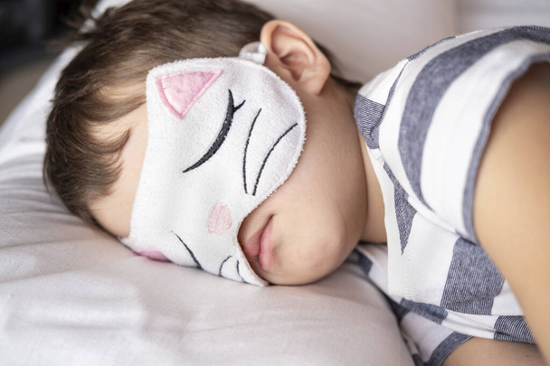  Милый дошкольник в маске для глаз котенка лежит в белой кровати.  - Фото, изображение