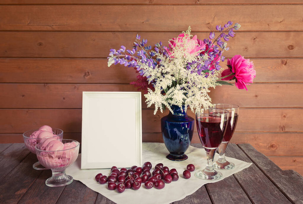 空の白いフレーム、青い花瓶の美しい花束、赤ワインのグラス、素朴な木製のテーブルの上にチェリーアイスクリームと新鮮なチェリーとガラスボウル。コピースペースで見る. - 写真・画像