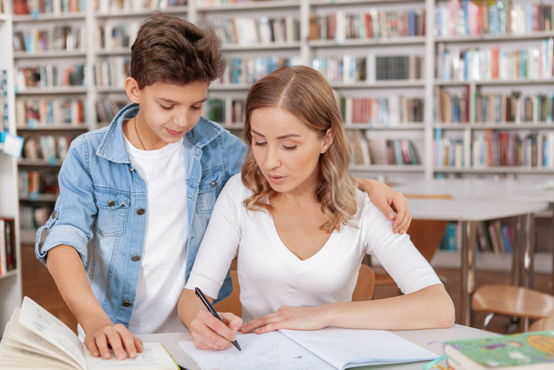 Attractive woman checking homework of her son at the library. Young boy studying with his mom - Φωτογραφία, εικόνα
