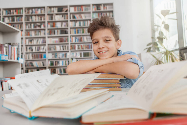 Lovely cheerful schoolboy smiling to the camera, sitting at the desk at the library - Photo, image