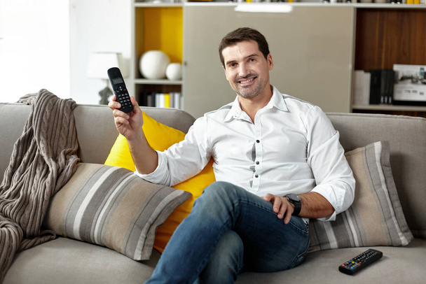 Ein lächelnder junger Mann sitzt gemütlich mit einem stationären Telefon auf der Couch und blickt in die Kamera. - Foto, Bild
