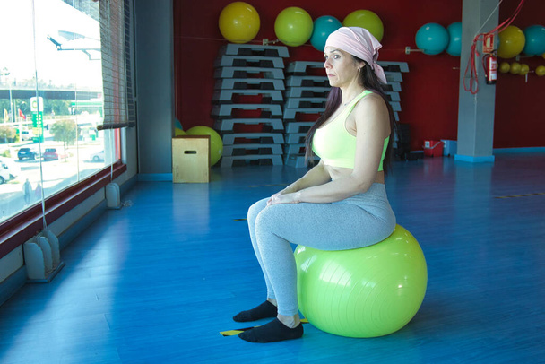 Mujer con bufanda rosa de cáncer sentada sobre una pelota de pilates amarilla con aspecto pensativo en el gimnasio, frente a una gran ventana y con suelo de parquet azul. - Foto, Imagen