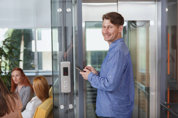 Cheerful businessman using smart phone while waiting for elevator at office building - Valokuva, kuva