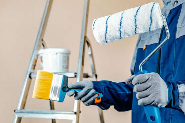 The plaster painter is ready to paint the wall. In the hands of a roller and brush. A stepladder and a bucket of paint in the background. - 写真・画像