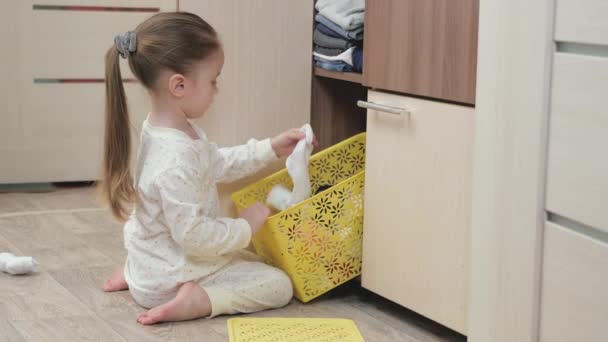 Ein Kind im Schlafanzug öffnet einen Schrank und sucht nach seiner Kleidung. Kleines Mädchen spielt im Kinderzimmer. Das Kind spielt herum und wirft seine Sachen aus der Box. Tochter spielt mit Kleidung zu Hause - Filmmaterial, Video