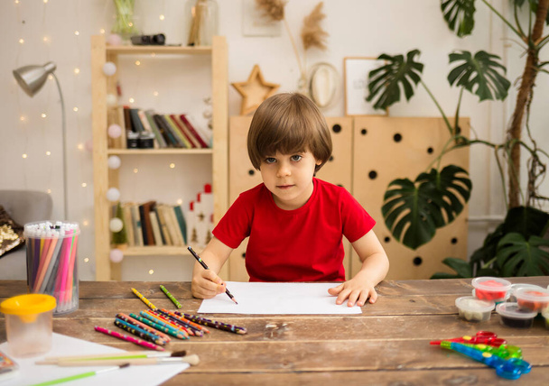 a small boy in a red T-shirt draws with a pencil on white paper at a wooden table and looks at the camera - Photo, Image