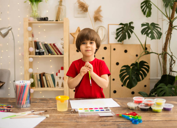 Kırmızı tişörtlü mutlu bir çocuk bir fırçayla çizer ve tahta bir masadaki beyaz kağıda resim yapar. - Fotoğraf, Görsel