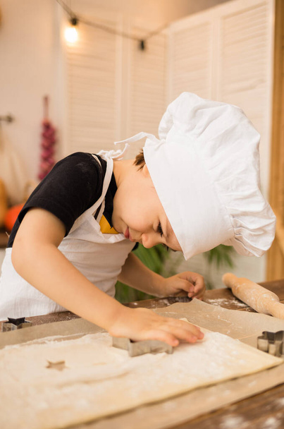 Ένας μικρός μάγειρας φτιάχνει μπισκότα από ζύμη σε ξύλινο τραπέζι στην κουζίνα. - Φωτογραφία, εικόνα