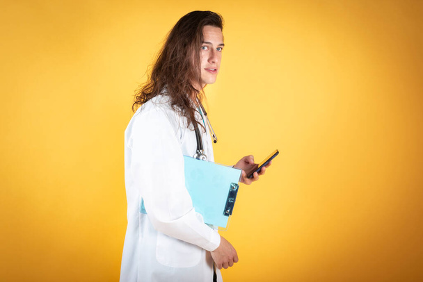 doctor with long hair, happy smiling, using smartphone on yellow background, copy space - Φωτογραφία, εικόνα