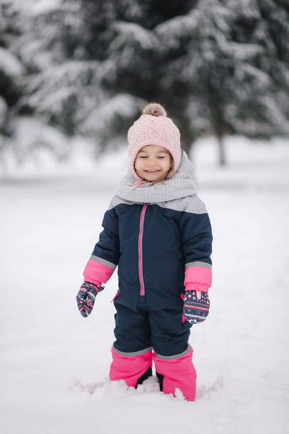 Boldog kislány sétál a parkban a hóban. Imádnivaló kislány rózsaszín kalapban és overálban, ahogy az erdőben sétál egy téli havas napon. - Fotó, kép