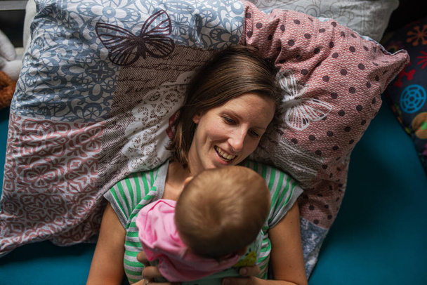 Jovem mãe feliz deitada na cama com sua menina em cima dela em um momento genuíno de alegria. - Foto, Imagem