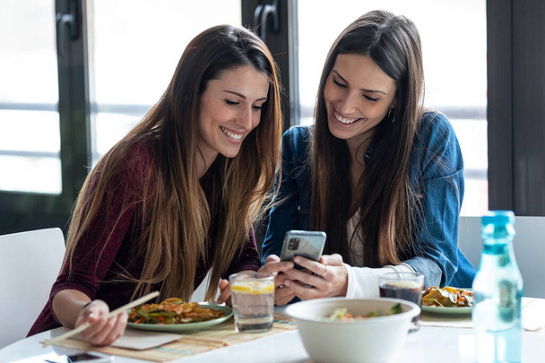 Shot of two beautiful women friends eating japan food while looking smart phone in the kitchen at home. - Fotografie, Obrázek
